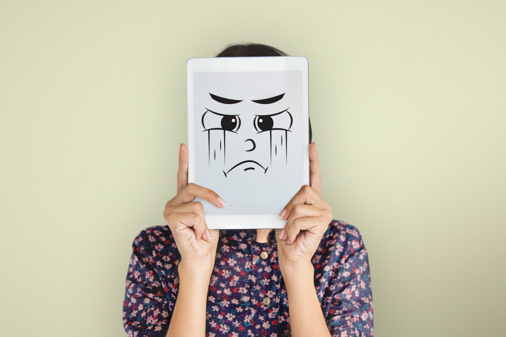 Woman holding a tablet displaying a cartoon crying and angry face, symbolizing hidden emotional turmoil and mood swings in bipolar disorder